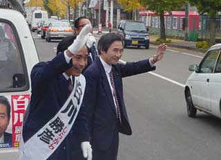 写真:道行く人々や車に手を振って支援をお願いする白川候補。右は長部登県議会議員